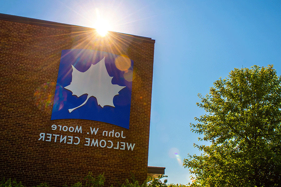 The sun is partially visible over the red brick wall of the John W. Moore Welcome Center. Rays of light shine over top the large white Sycamore leaf on the blue background that appears on the side of the building with the name underneath. A green tree is also visible to the right against the blue sky. 