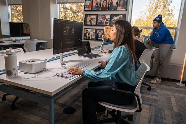 A female student sits in front of a large computer monitor while editing a video. She has dark hair and wears an aqua-blue blouse and dark blue slacks. In the background, another female student is partially visible seated in front of a monitor, while a male student in a blue hoodie and black-and-blue winter cap looks over her shoulder. A laptop sits on the table between the two women. 