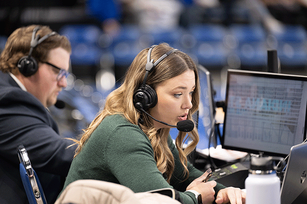 A female student with blondish-brown hair and wearing a green sweater is seated in a sports stadium, with rows of blue seats visibly blurred behind her. She is wearing headphones and a mic, holds a pen in her hand, and is looking at laptop and desktop computer monitors that are set up in front of her. A male student with brown hair and glasses, who is wearing a grey suit jacket and white collared shirt, is slightly blurred but visible behind her. He is wearing headphones.